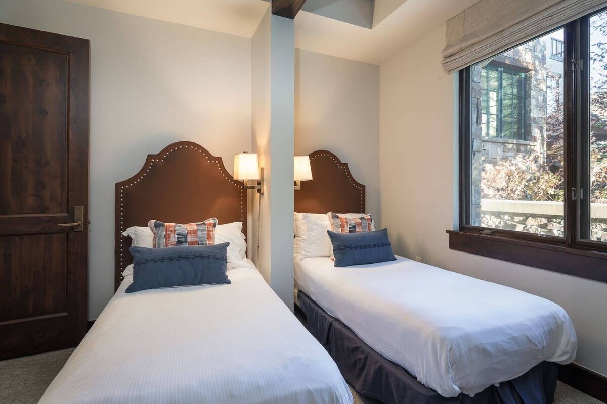 Two single beds with wooden headboards and bedside lamps, a wooden wardrobe, and a window with a view of trees.