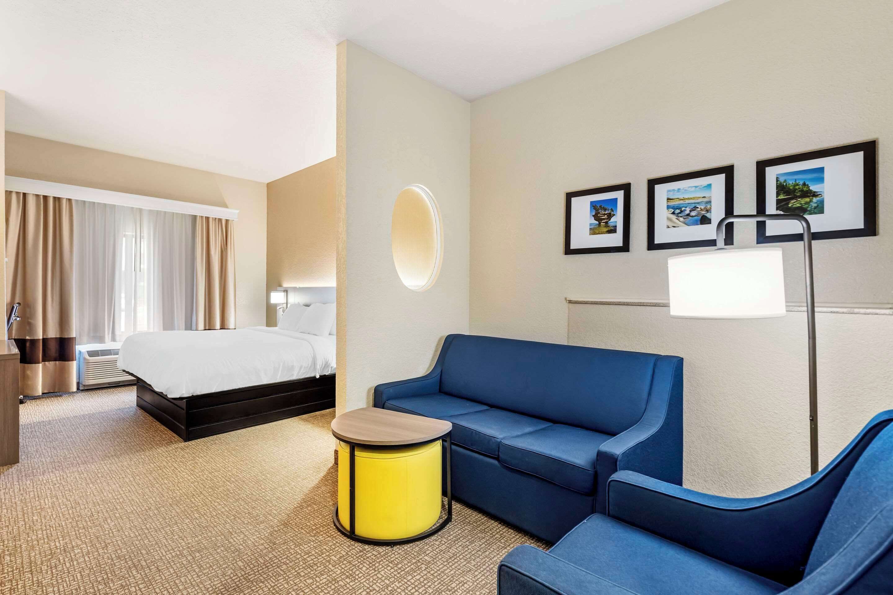 A hotel room with a bed, a blue sofa, a small yellow table, and a floor lamp. There are three framed pictures on the wall.