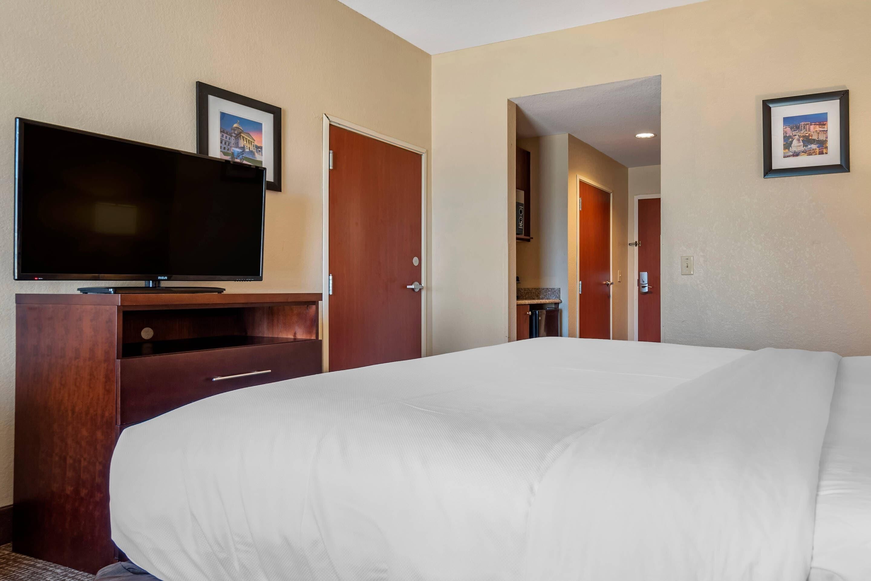 Une chambre d’hôtel avec un grand lit, un téléviseur à écran plat posé sur un meuble en bois, une porte donnant accès à une salle de bain et un tableau encadré accroché au mur.