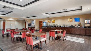 A hotel dining area with multiple tables and chairs, a kitchen area with a coffee station, and a trash bin.