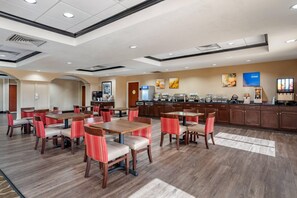 A hotel dining area with multiple tables and chairs, a kitchen area with a coffee station, and a trash bin.