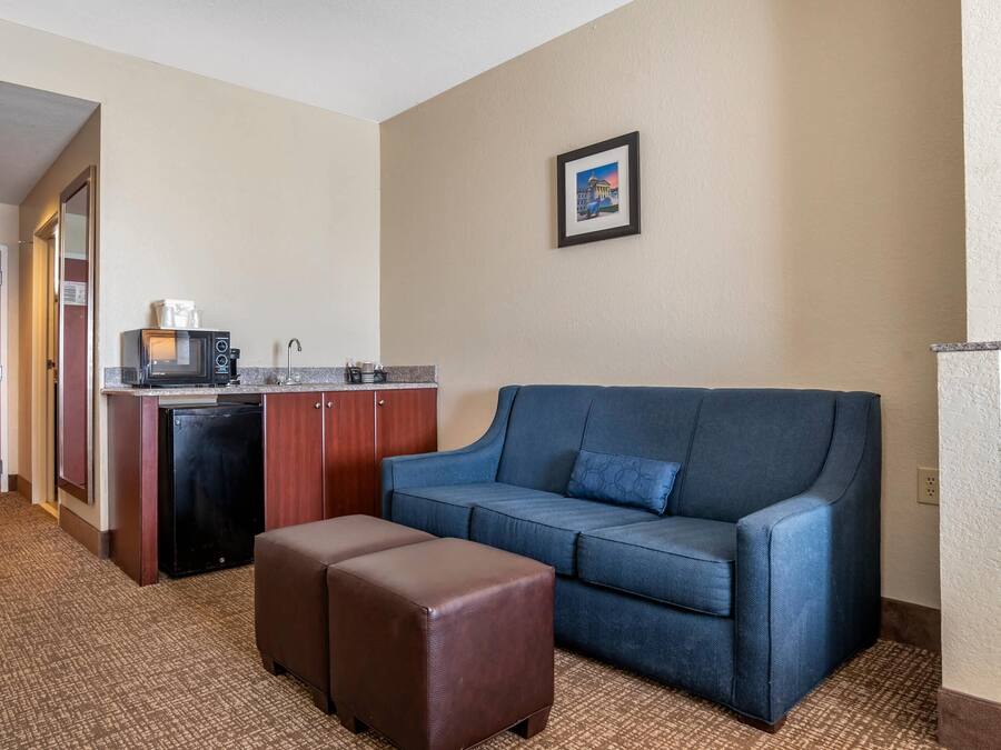 A compact hotel room with a kitchenette, a blue sofa, a brown ottoman, and a framed picture on the wall.