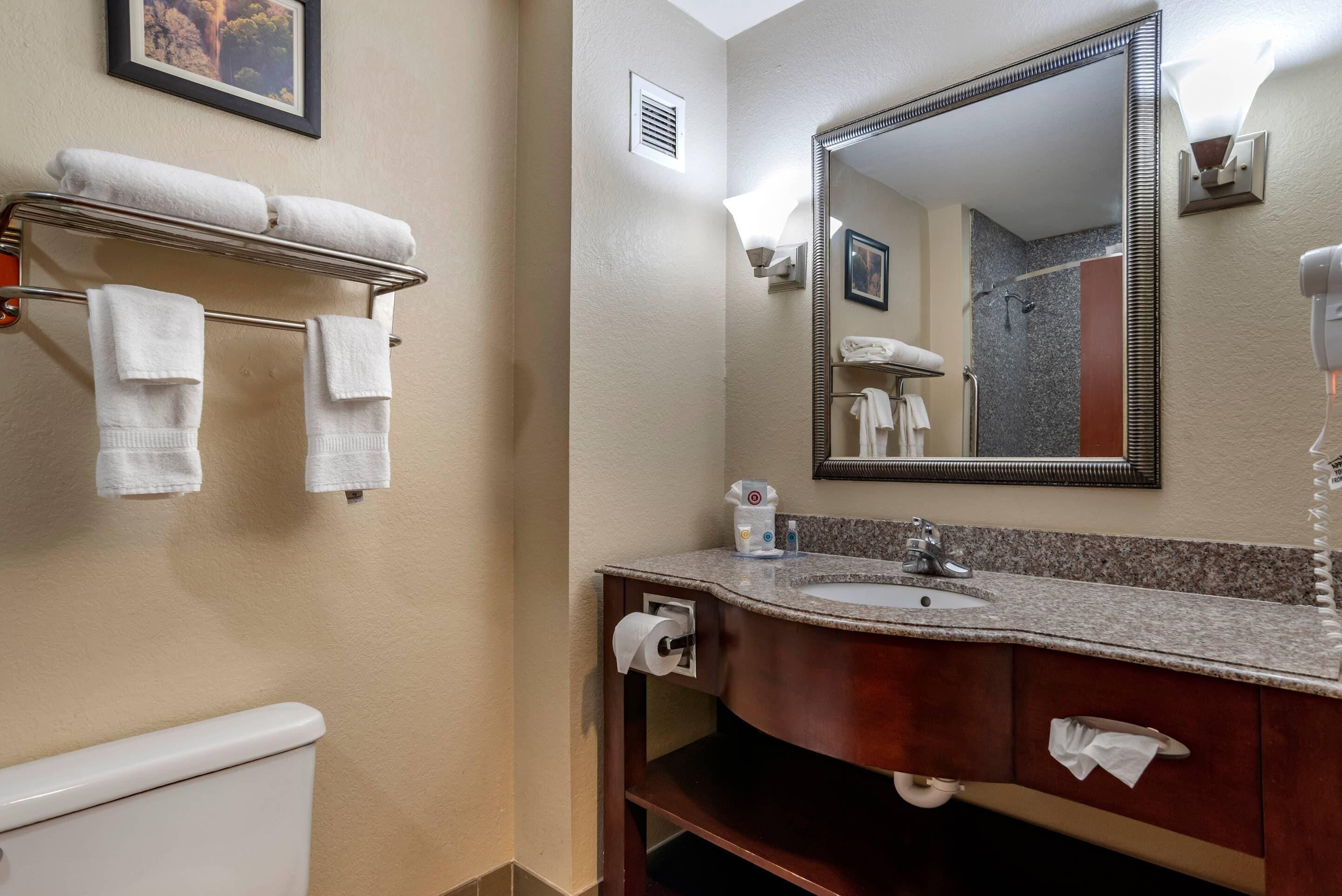 A bathroom with a toilet, a sink with a mirror, towels on a rack, and a towel dispenser.