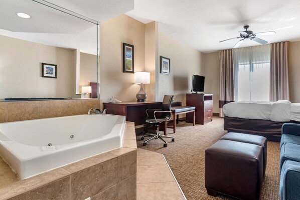 A hotel room with a large bathtub, a desk with a chair, a bed, a ceiling fan, and a television.