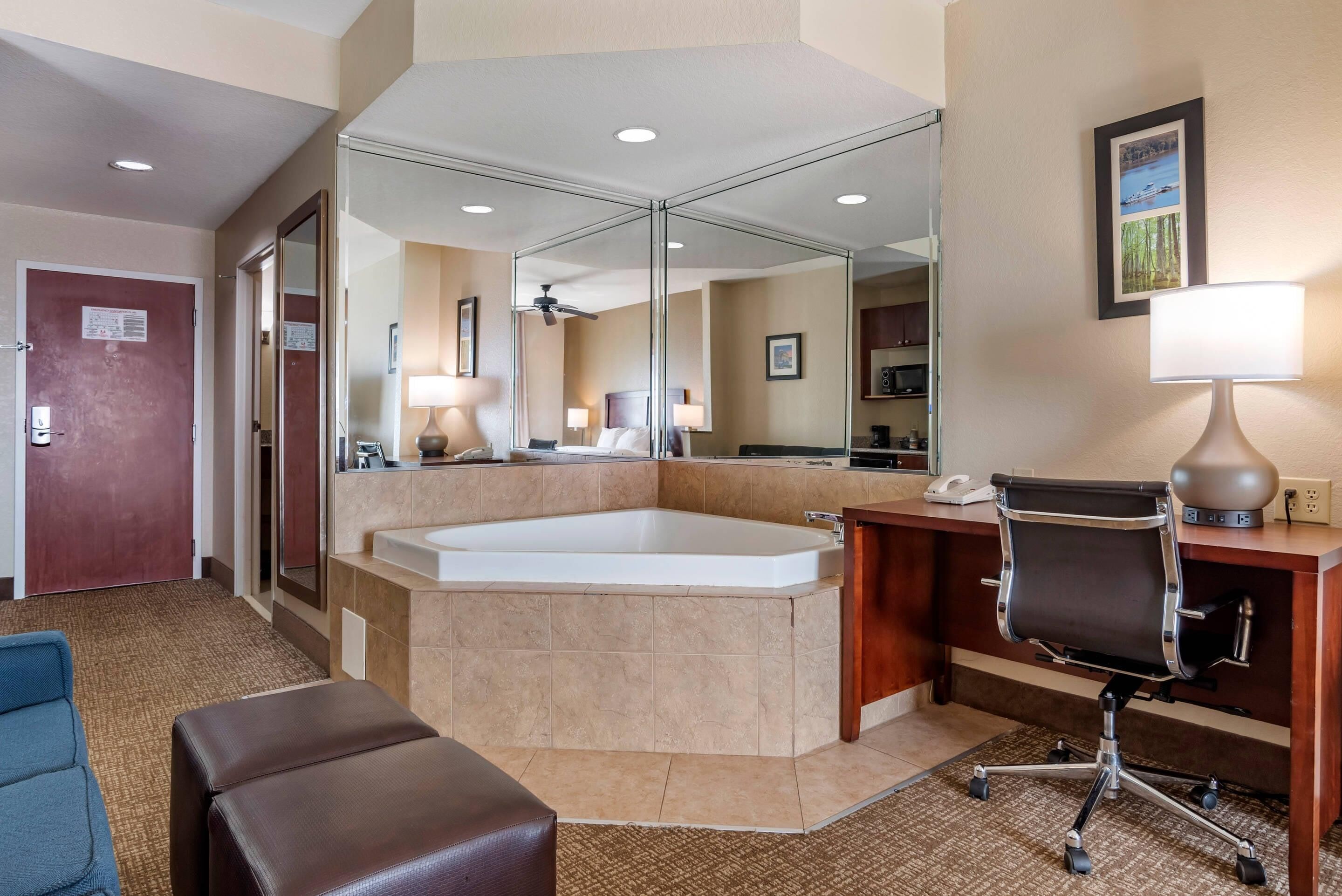 A hotel room with a large bathtub, a desk with a chair, a mirror, and a wall-mounted lamp.