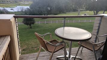 Um deck de madeira com uma mesa redonda e cadeiras de vime, com vista para uma área gramada com árvores e um corpo d'água.
