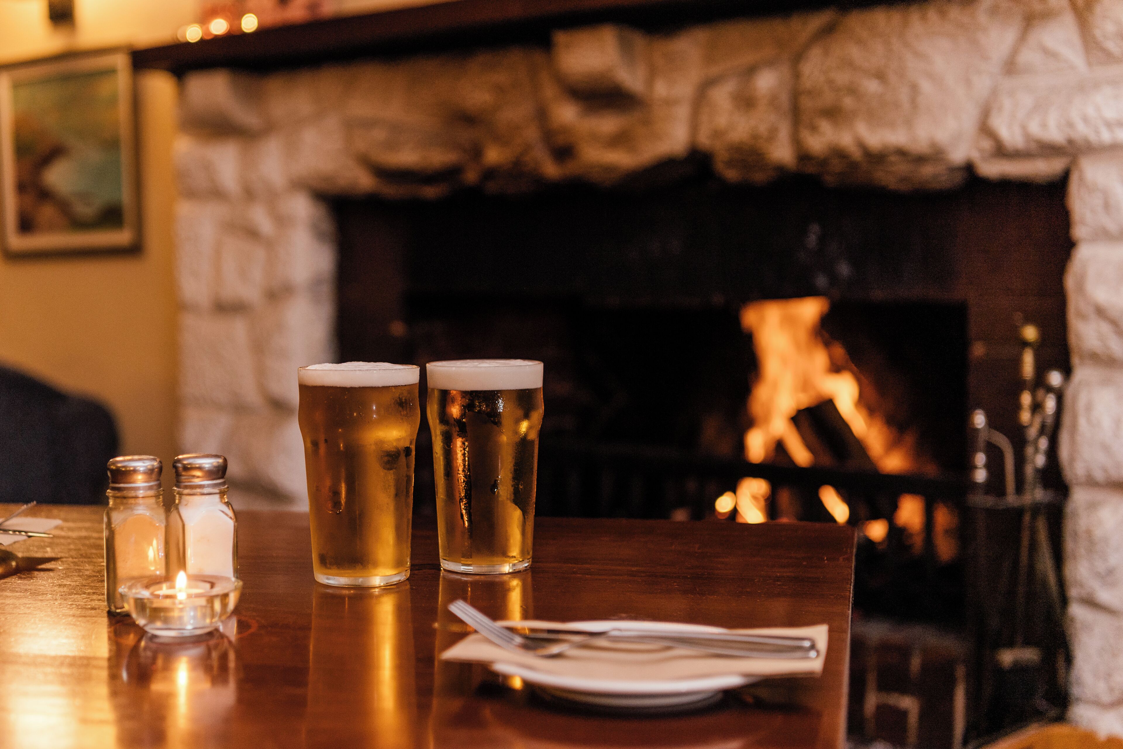 A table with two glasses of beer, salt and pepper shakers, and a plate with cutlery. A fireplace is visible in the background.