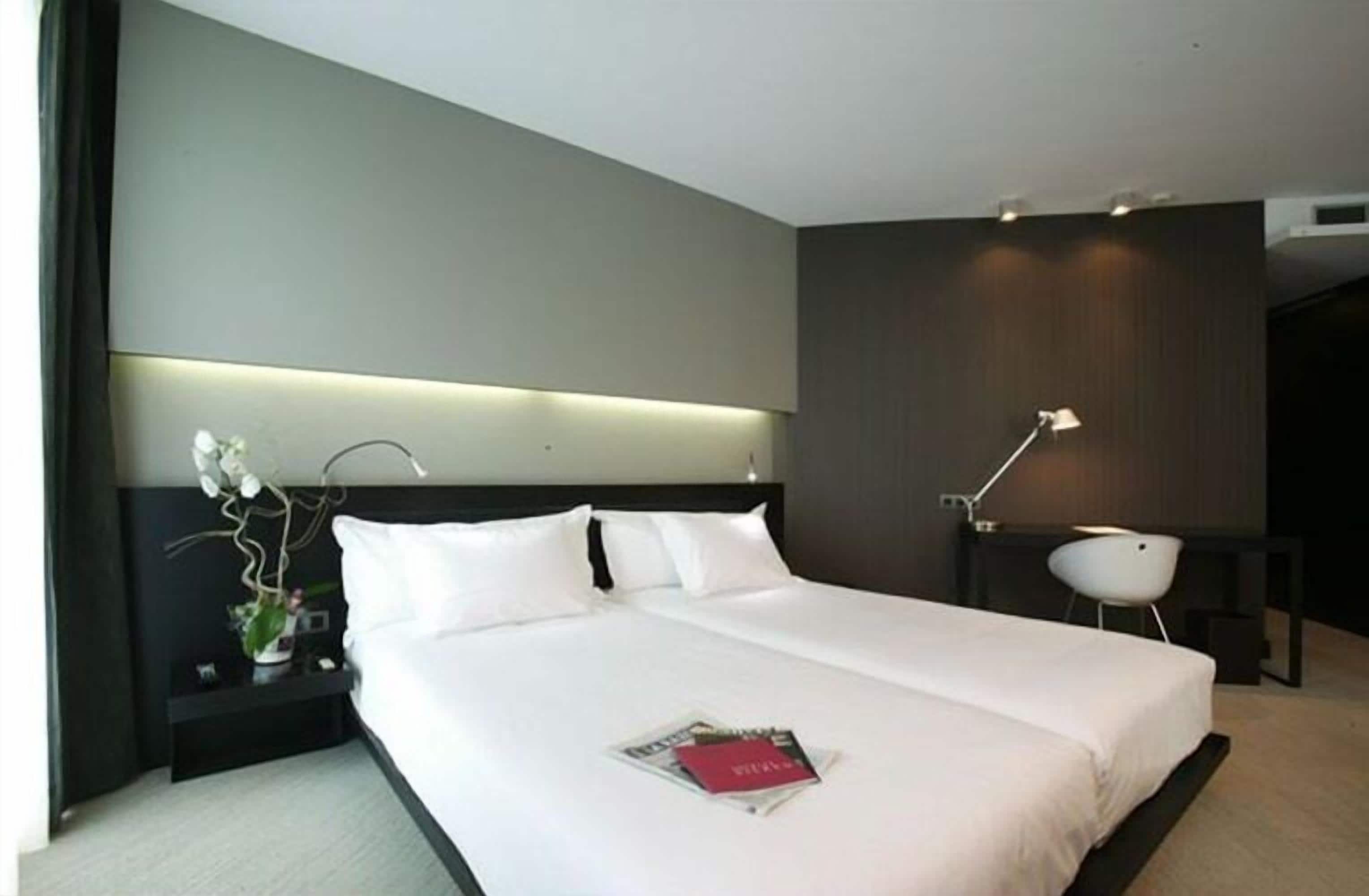 A modern hotel room with a large bed, a bedside table with a lamp, and a small desk with a chair.