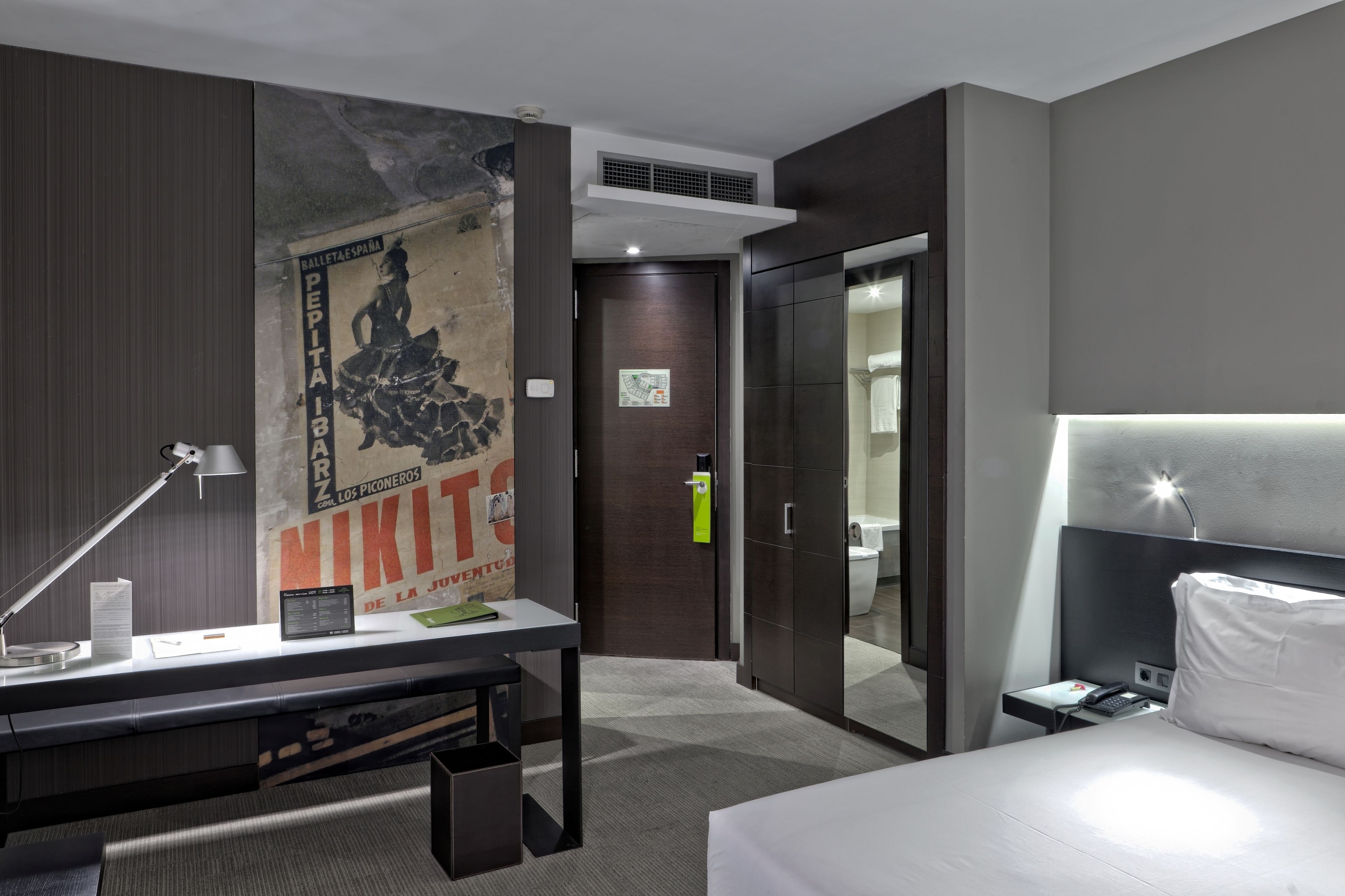 A modern hotel room with a large bed, a desk with a lamp, a poster on the wall, and a wardrobe.