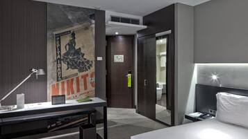 A modern hotel room with a large bed, a desk with a lamp, a poster on the wall, and a wardrobe.