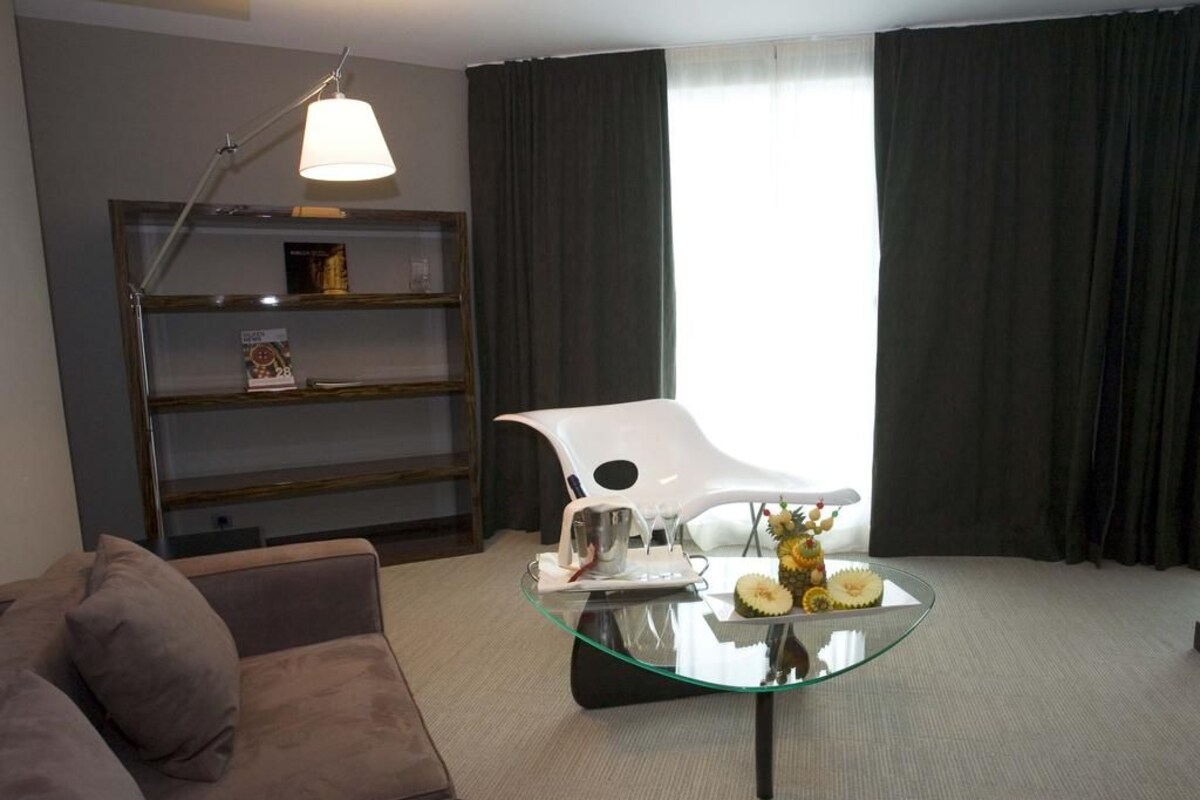 Una sala moderna con una mesa de centro de vidrio, un sillón blanco, un sofá, una estantería y una lámpara.