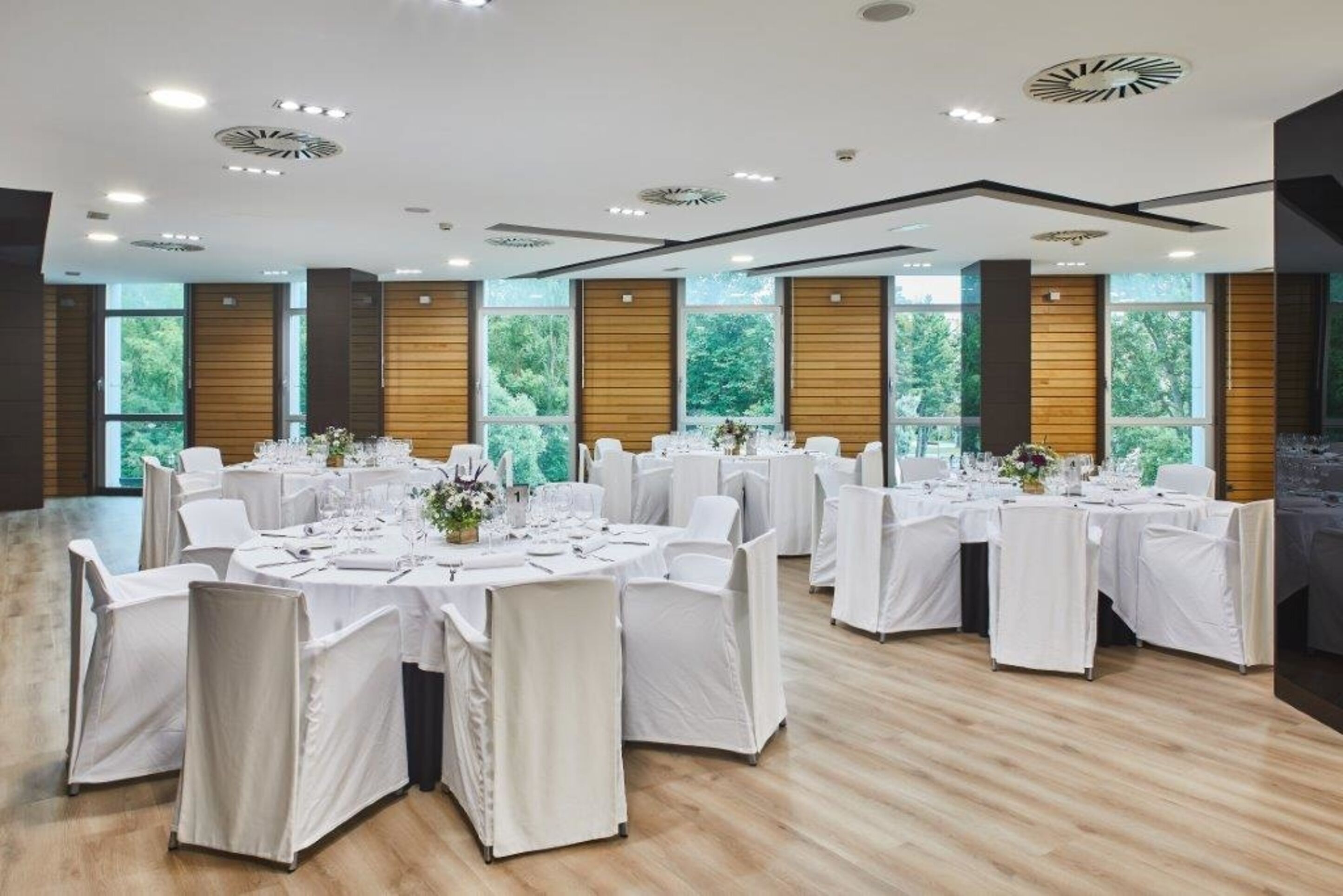 A conference room with multiple tables set for a formal event, large windows with a view of greenery, and a modern ceiling design.