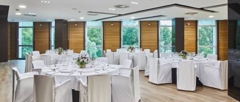 Un salón de conferencias con varias mesas dispuestas para un evento formal, grandes ventanales con vistas a zonas verdes y un diseño moderno de techo.