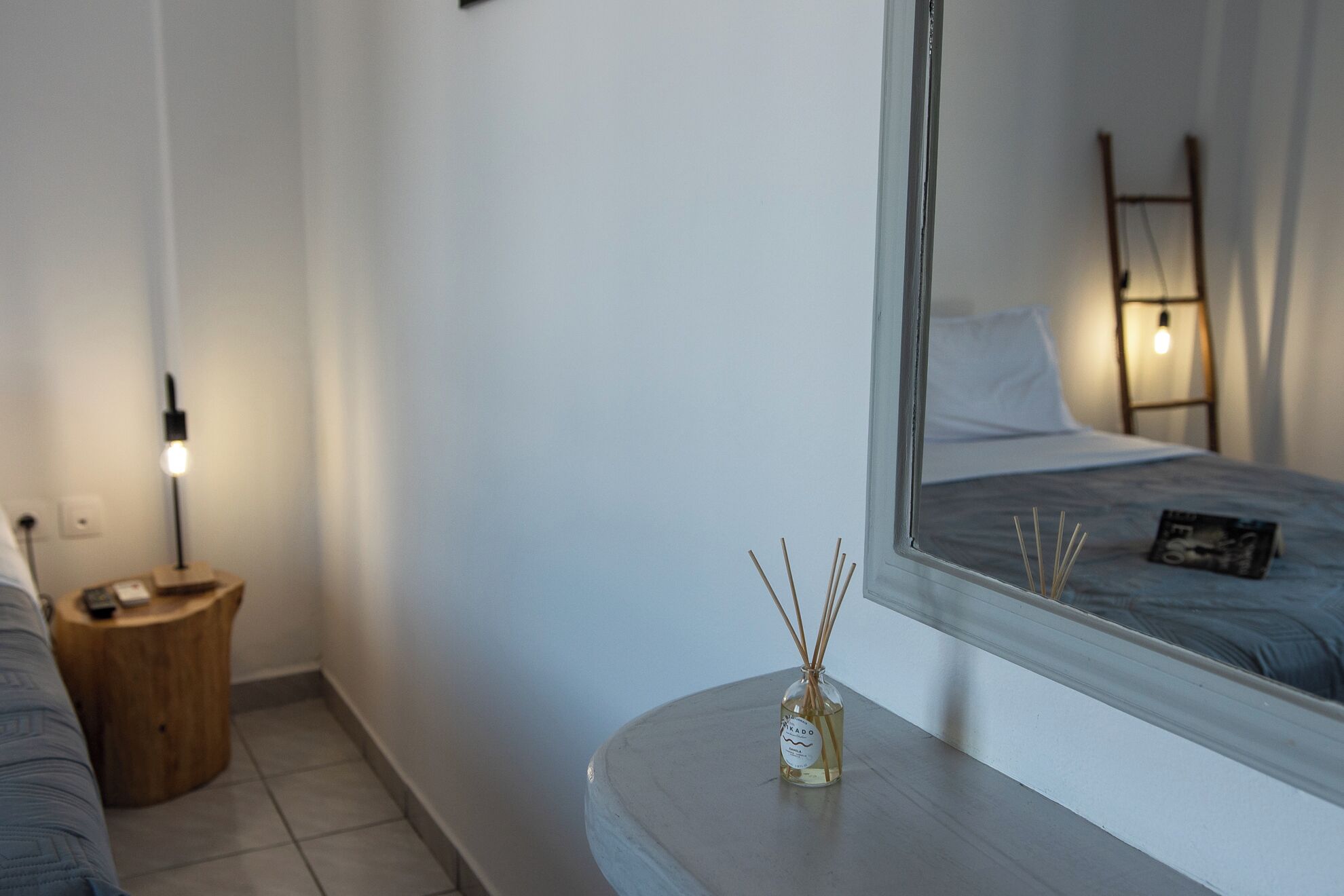 A bedroom with a bed, a bedside table with a bottle of reed diffuser, a bedside lamp, and a mirror reflecting the bed and lamp.