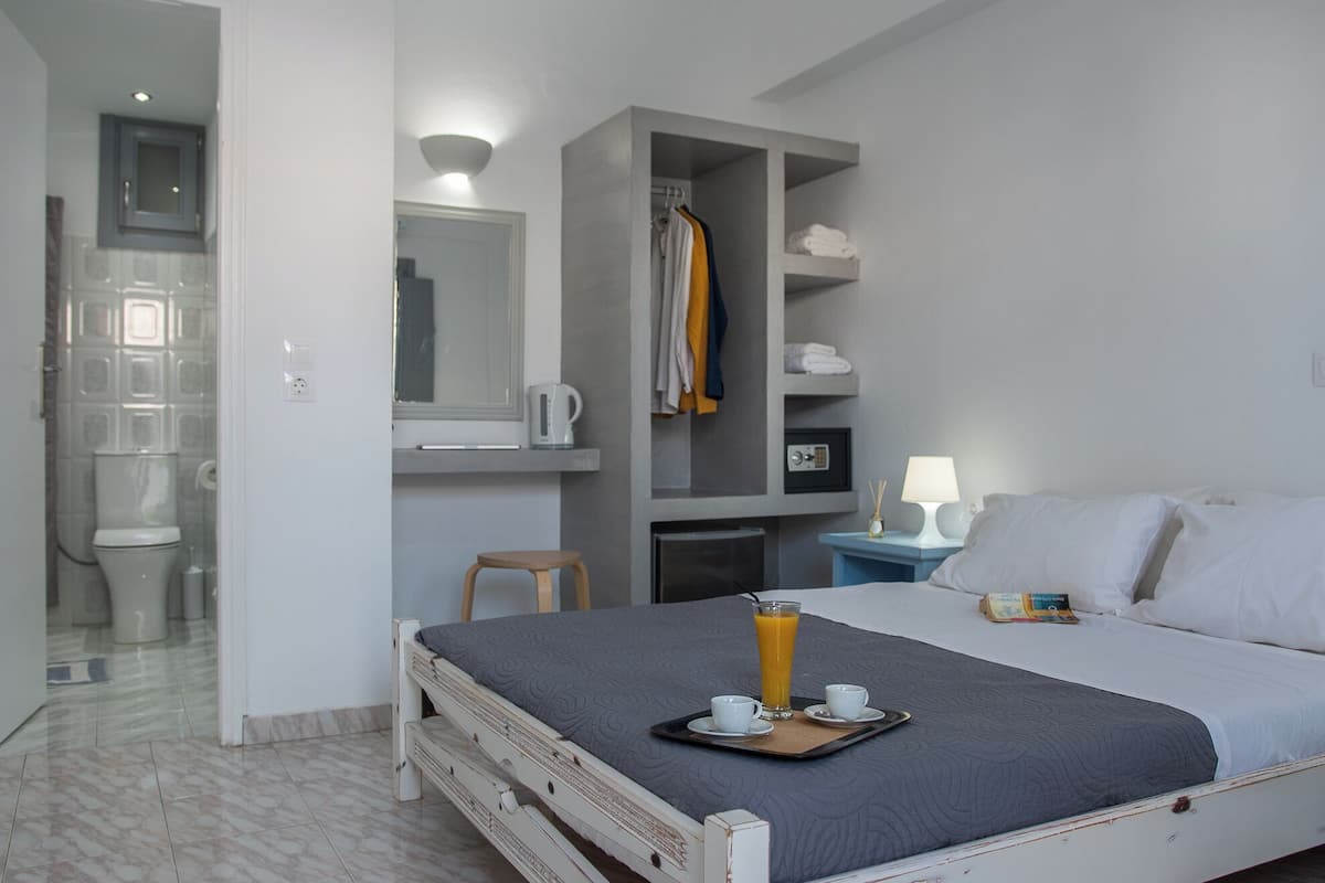 Quarto de hotel com cama, mesa de cabeceira com abajur, um banquinho, prateleira com toalhas e banheiro com vaso sanitário e chuveiro.