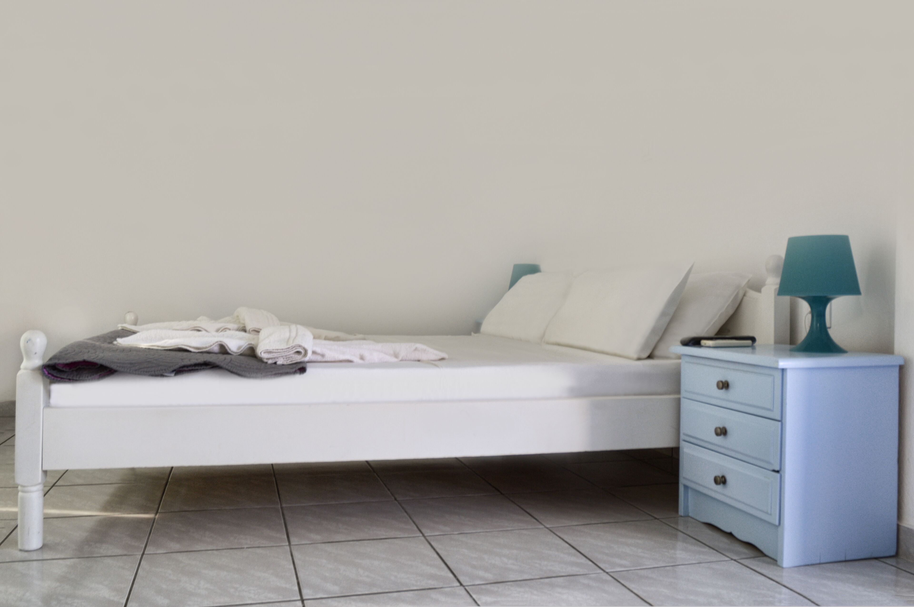Una cama bien hecha con almohadas y una manta, una mesita de noche con lámpara y un tocador blanco con cajones.