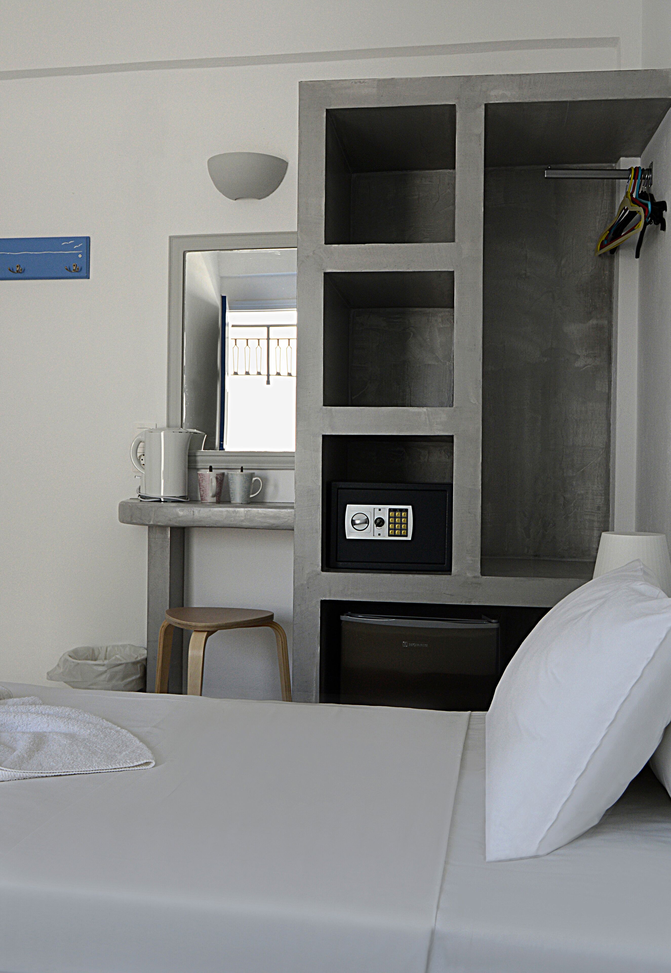 A modern hotel room with a bed, a small stool, a coffee station, a safe, and a mirror.