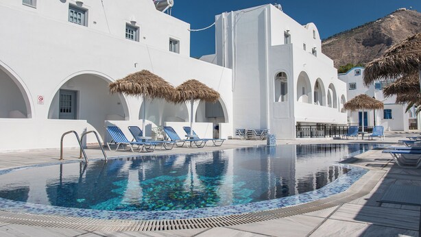 Un espace piscine doté de chaises longues et de parasols, entouré de bâtiments blancs ornés d’arches, avec une montagne en toile de fond.