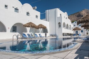 Uma área de piscina com espreguiçadeiras e guarda-sóis, cercada por edifícios brancos com arcos e uma montanha ao fundo.