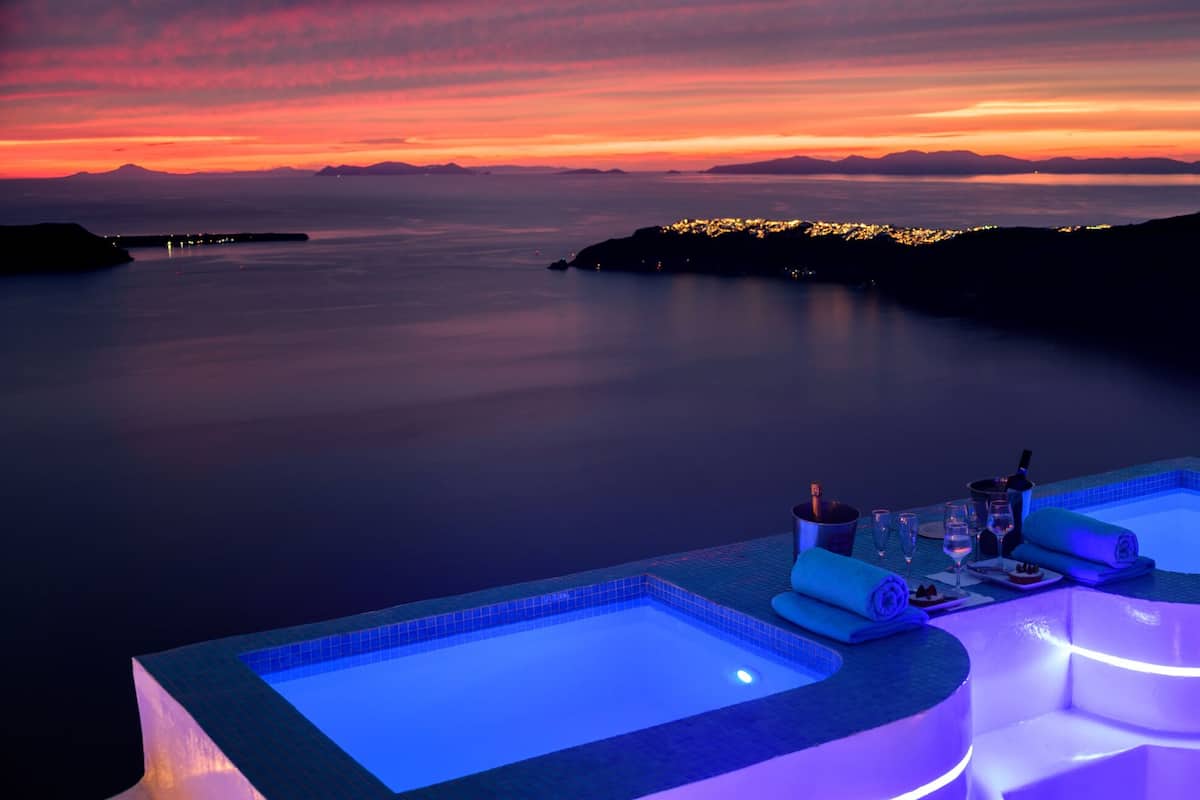 A luxurious pool area with two hot tubs, a dining setup, and a view of the sea at sunset.