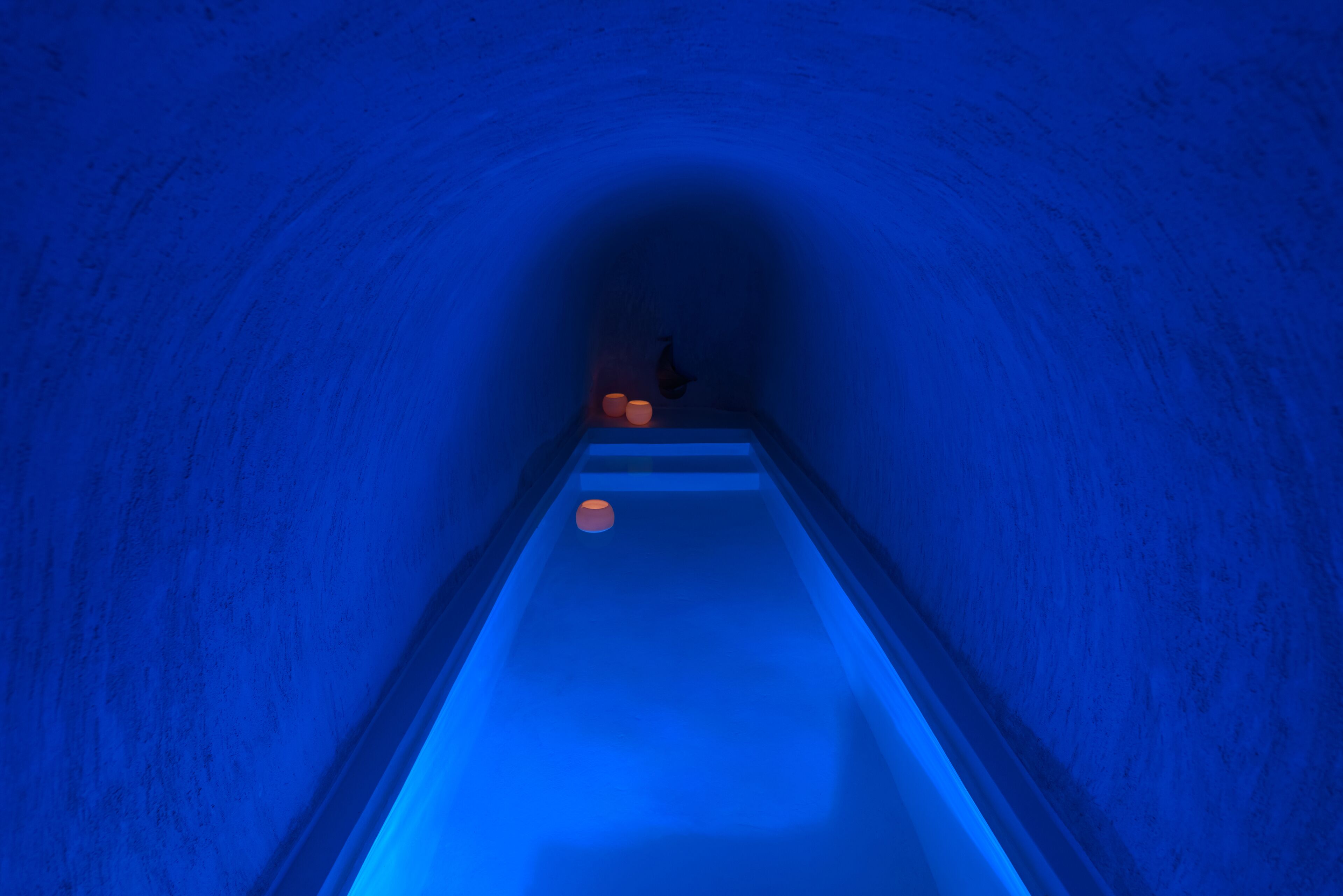 A narrow, blue-lit tunnel with a person walking towards a platform with orange objects.