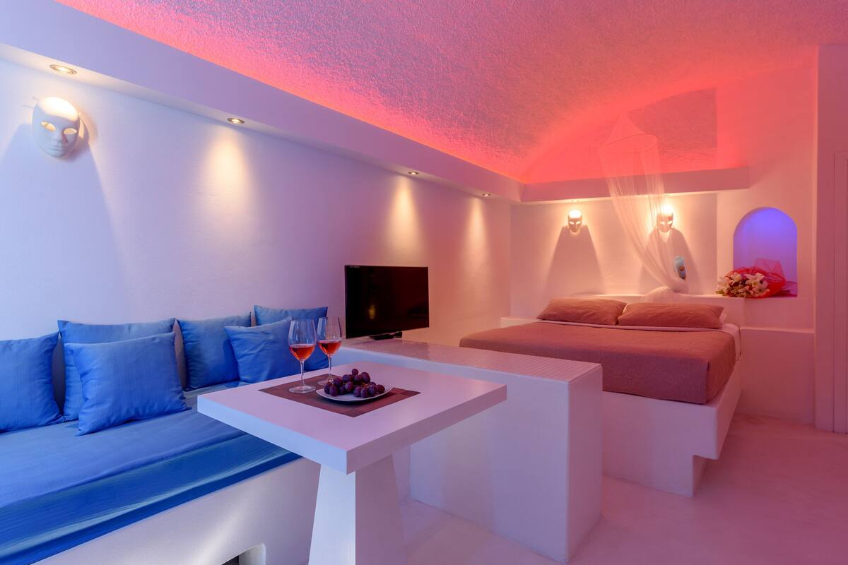 A modern bedroom with a sofa, a bed, a TV, and a table with a fruit bowl and glasses.