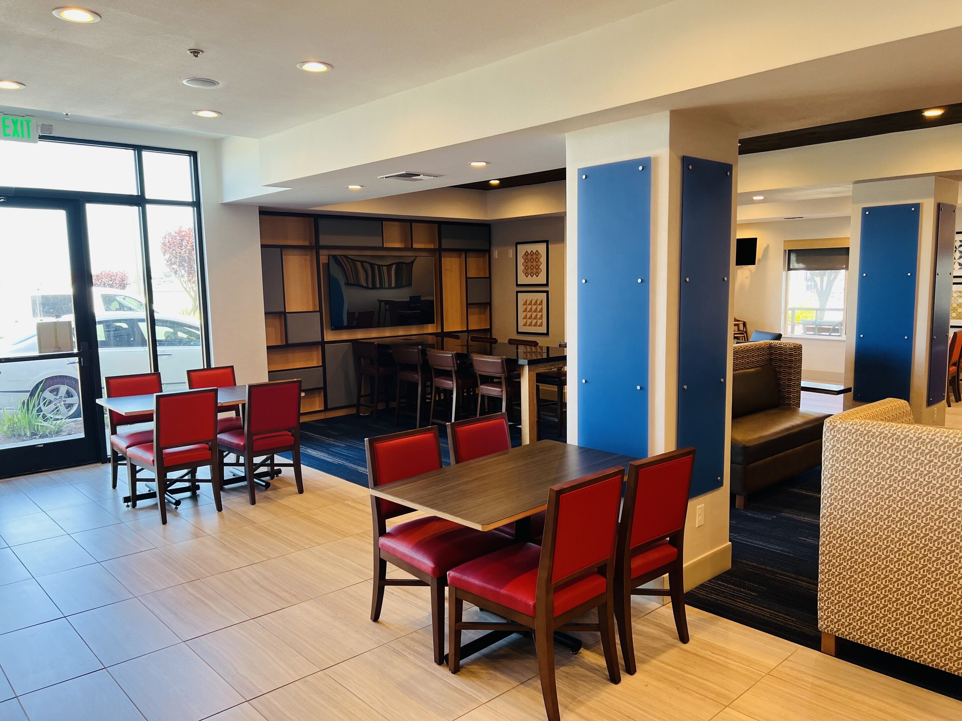A restaurant with a large dining area featuring red chairs and a wooden table. There are blue and white pillars, and a kitchen area in the background.