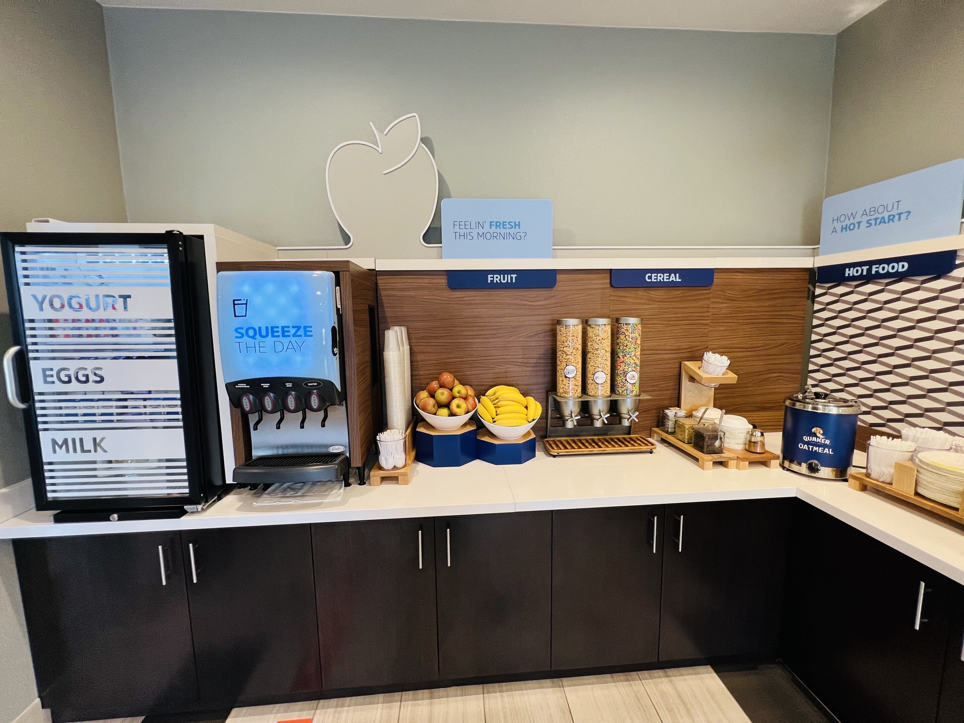 A breakfast bar with yogurt, eggs, milk, and a squeeze station. There are bowls of fruit and cereal, and a hot food station.