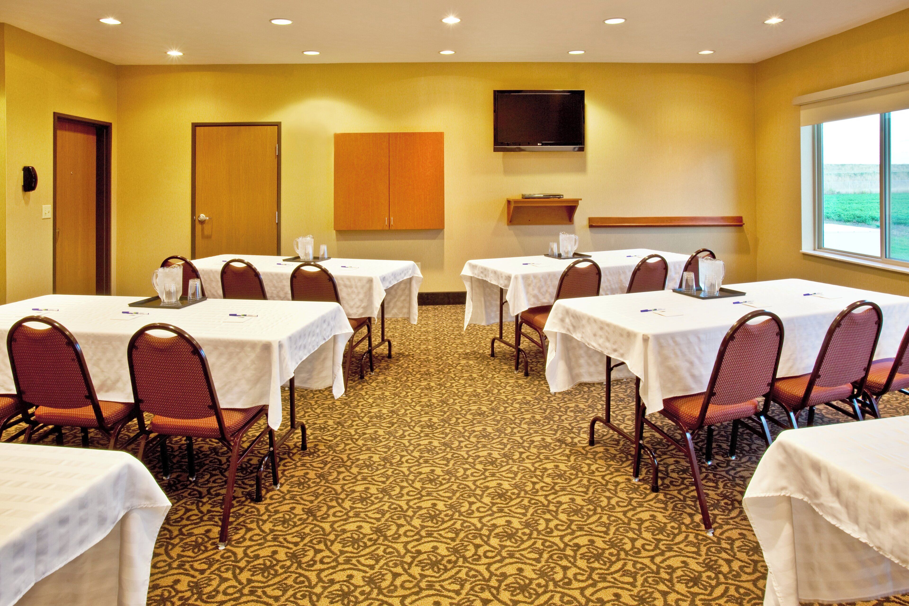 A conference room with multiple tables and chairs, a flat-screen TV mounted on the wall, and a window providing a view of the outdoors.