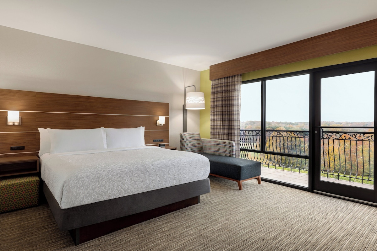 Habitación de hotel con una cama grande, una silla, una mesita de noche, una lámpara y un balcón con vistas.