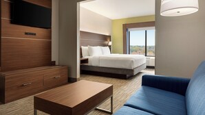 A modern hotel room with a flat-screen TV mounted on a wooden panel, a bed with white bedding, a blue sofa, and a large window offering a view of the outdoors.