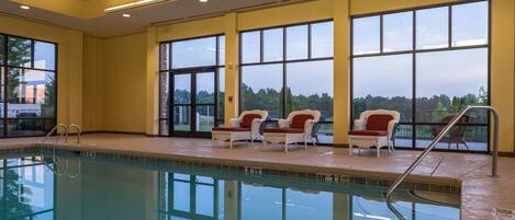 An indoor swimming pool with clear glass walls, a wooden ceiling, and a view of trees outside.