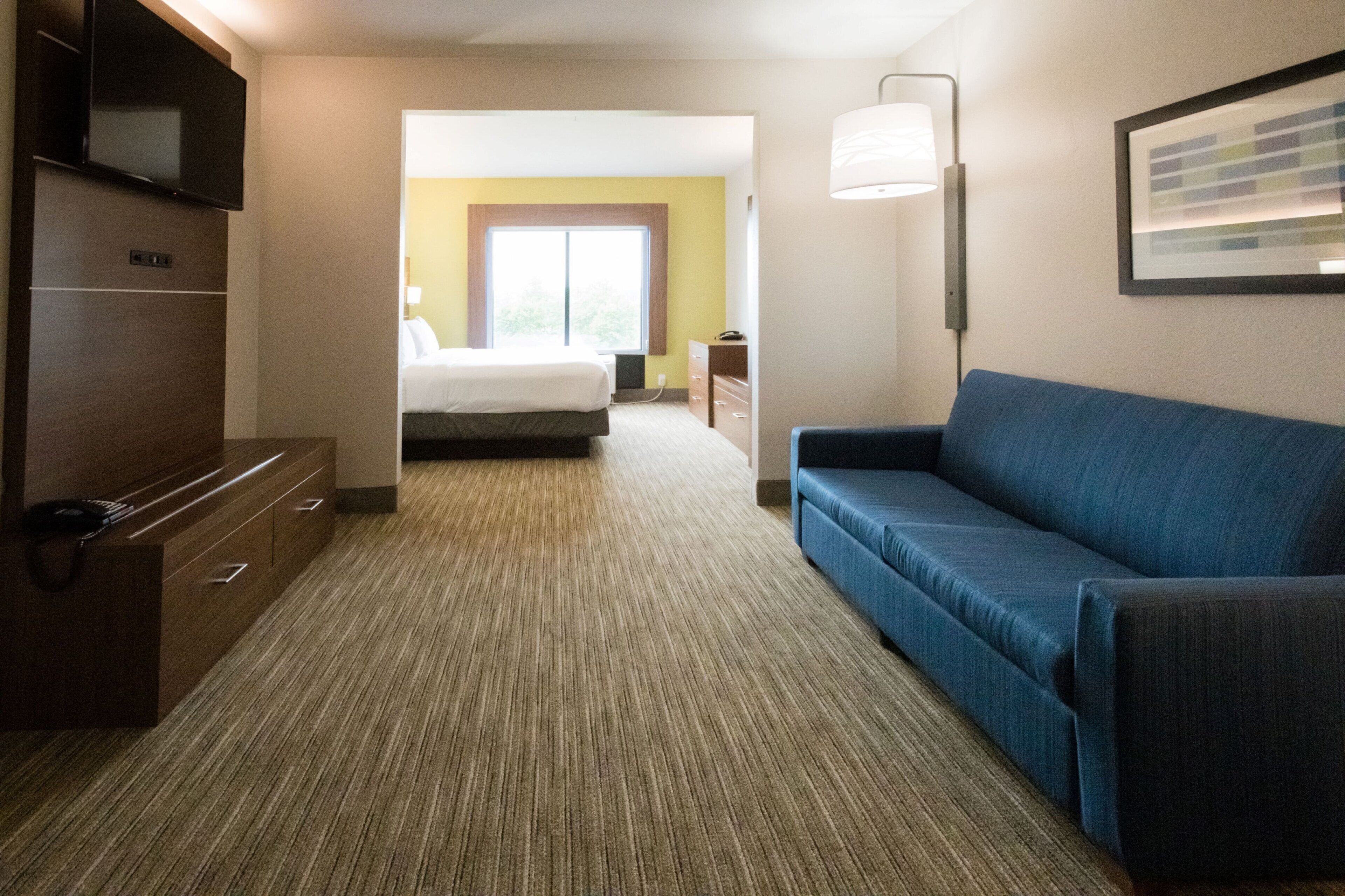 A hotel room with a bed, a blue sofa, a TV, a desk with drawers, and a lamp.