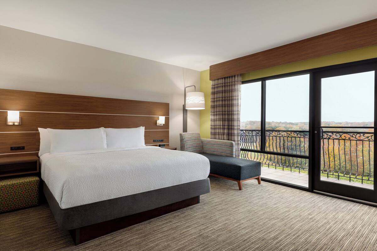 A hotel room with a large bed, a chair, a nightstand, a lamp, and a balcony with a view.