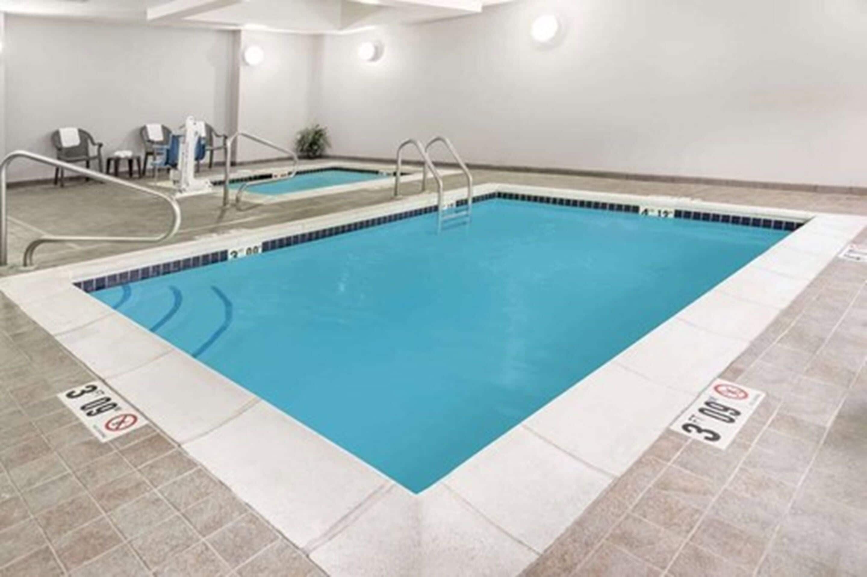 An indoor swimming pool with clear lane markings, a lifeguard chair, and a small seating area with chairs.