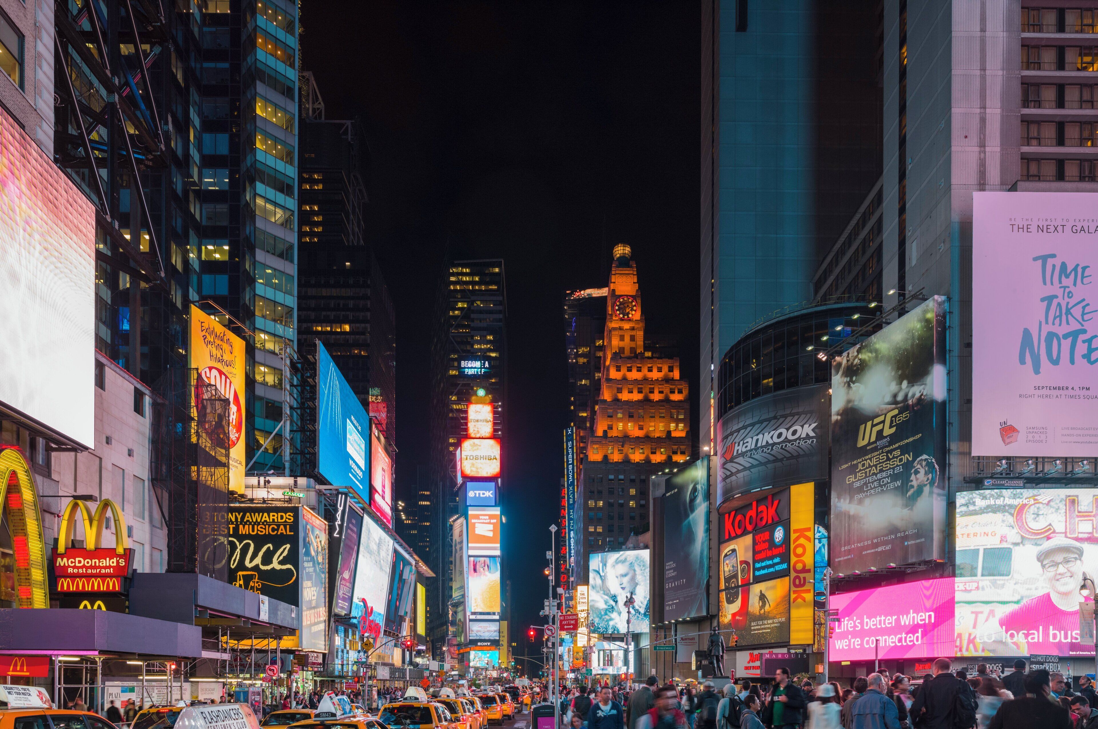 Uma rua urbana movimentada à noite, com outdoors e placas iluminadas.