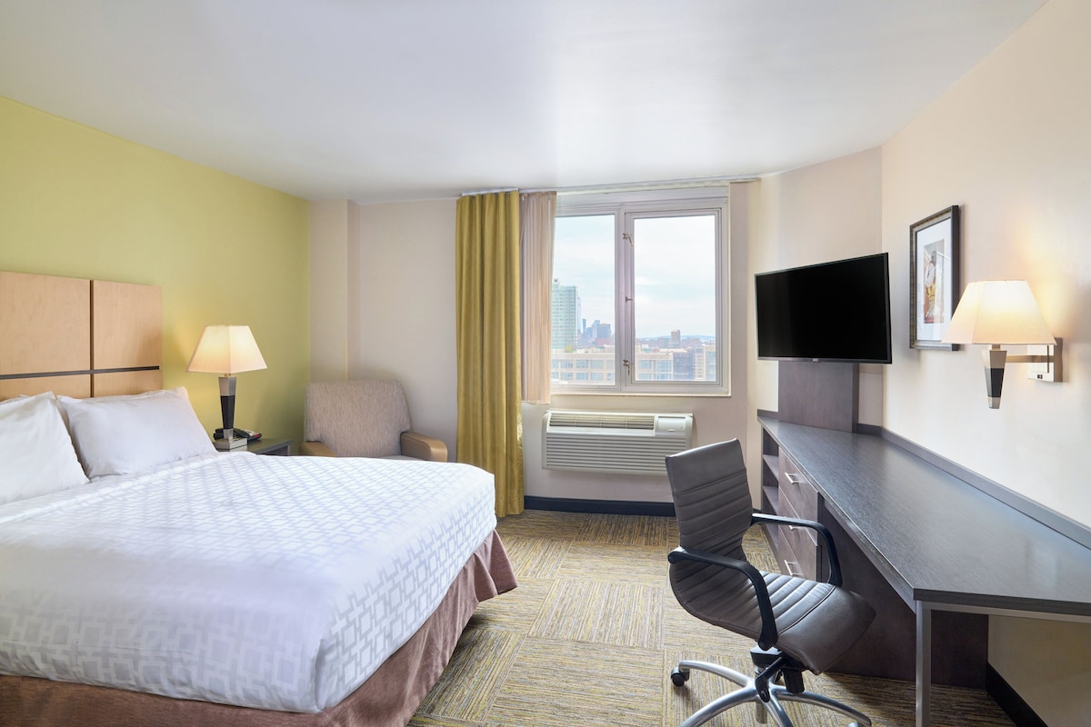 Habitación de hotel con cama, escritorio, silla, televisor y ventana con vista a la ciudad.