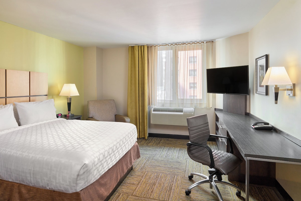 Una habitación de hotel con cama, escritorio, silla, televisor y aire acondicionado.