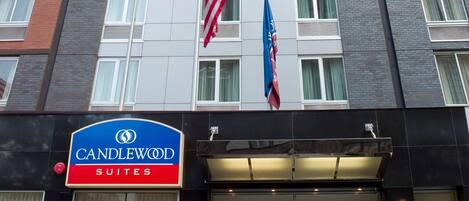 Entrada de un hotel con un cartel de Candlewood Suites, dos plantas en macetas y dos banderas.