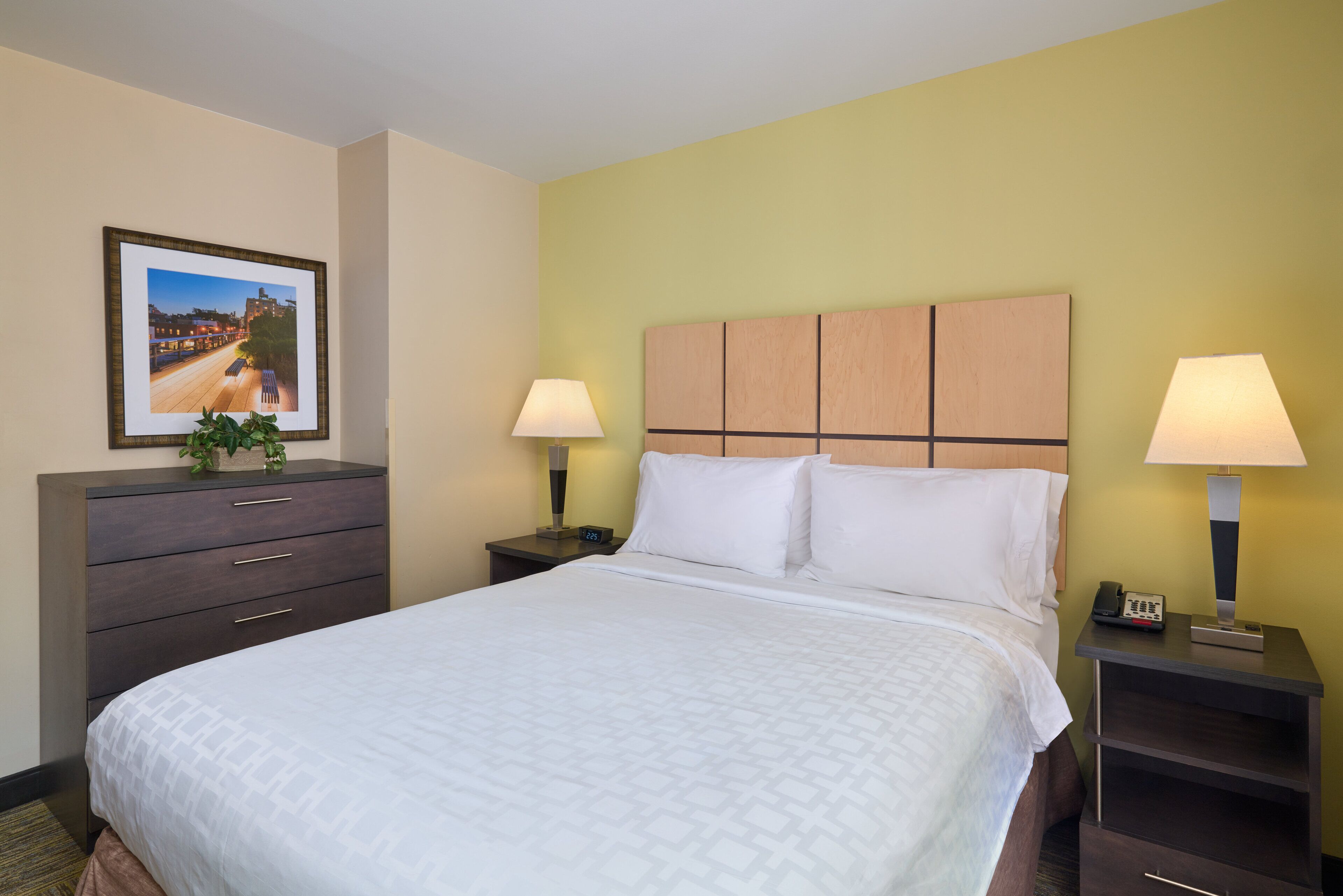 Habitación de hotel con una cama grande, dos mesitas de noche, un tocador, un aparador, un cuadro enmarcado en la pared y una planta en maceta.