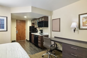 Habitación de hotel con cama, escritorio, silla, microondas, cafetera y un reloj en la pared.