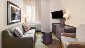 Habitación de hotel con sofá, una silla, una mesita, televisor de pantalla plana, lámpara, cuadro enmarco y ventana con cortinas.