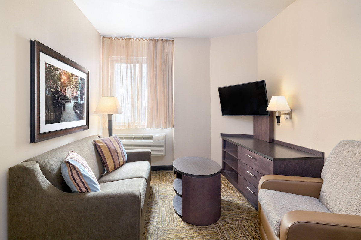 Habitación de hotel con sofá, una silla, una mesita, televisor de pantalla plana, lámpara, cuadro enmarco y ventana con cortinas.