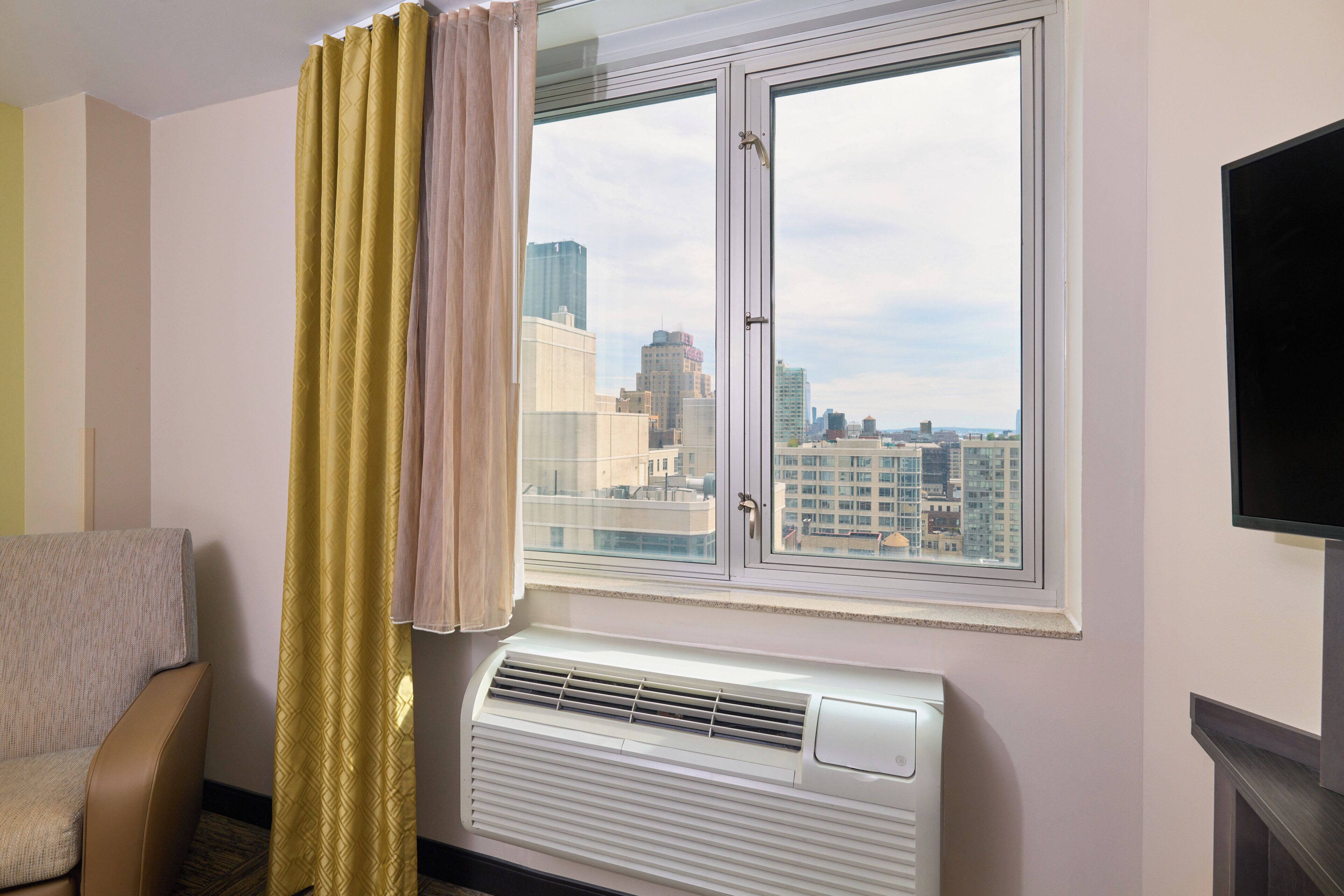 Ein Hotelzimmer mit großem Fenster, Klimaanlage und Blick auf eine Stadtlandschaft.