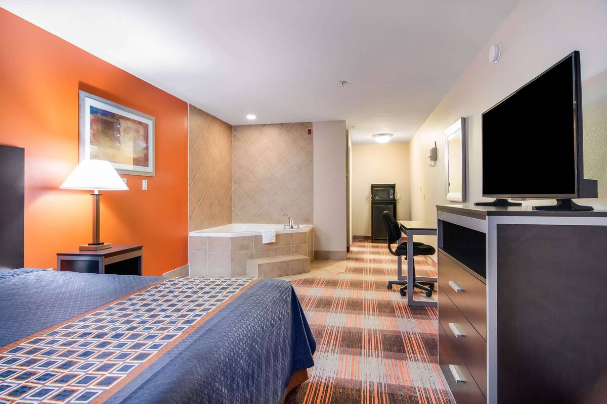 A hotel room with a bed, a desk, a television, a bathroom with a bathtub, and a painting on the wall.