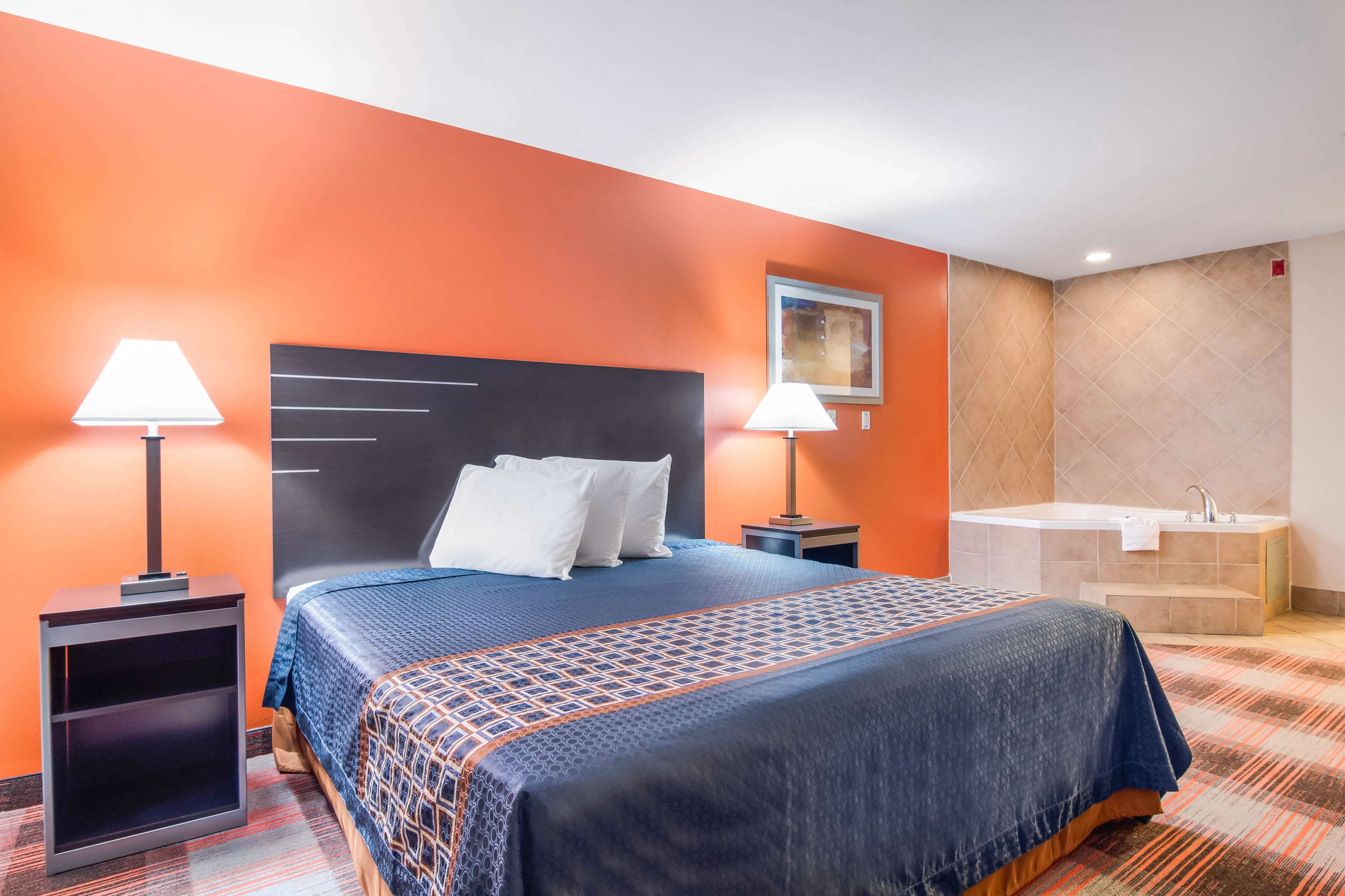 Une chambre d’hôtel avec un grand lit, deux lampes de chevet, une tête de lit, un tableau, un mur orange, une salle de bain avec une baignoire et un lavabo.