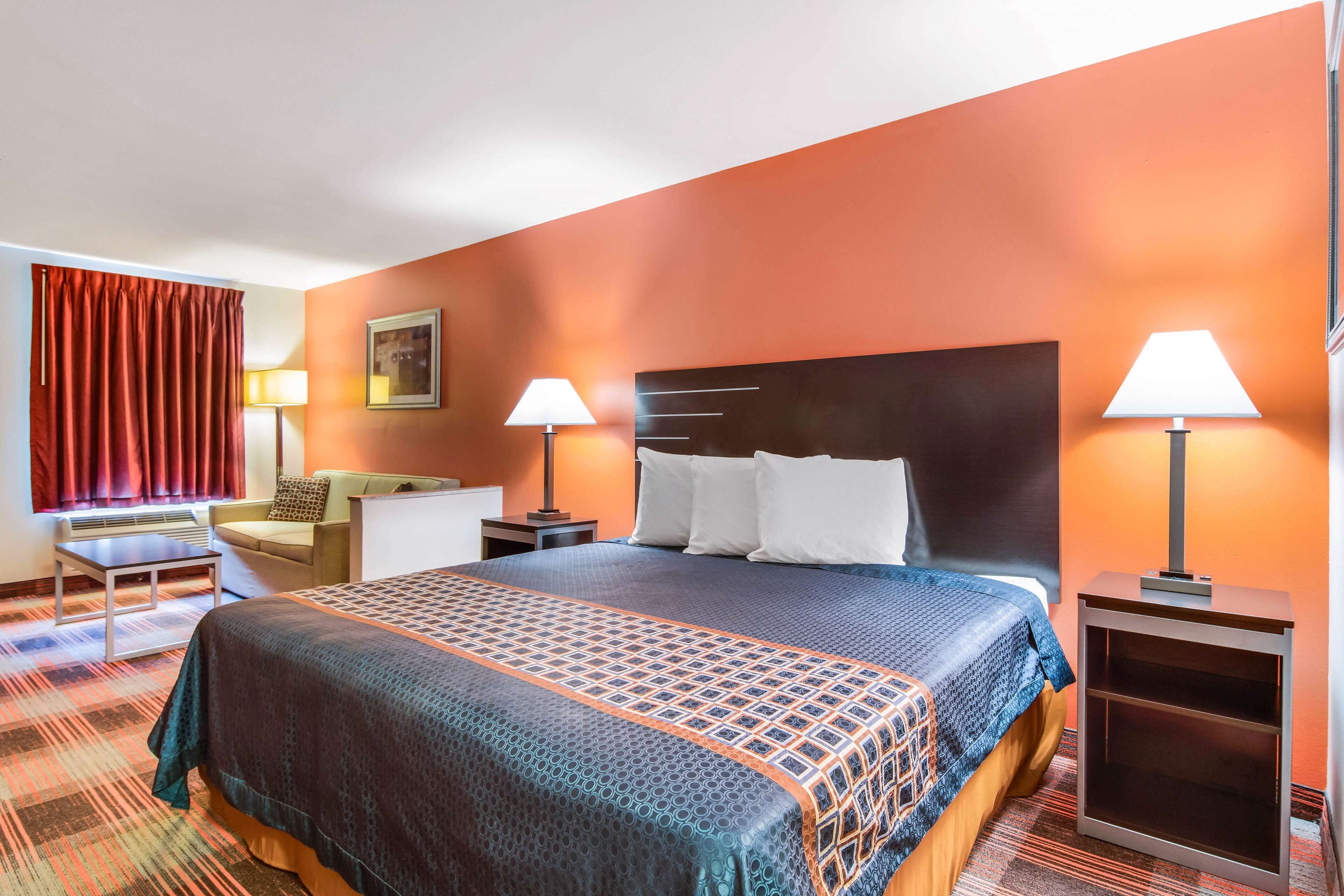 Habitación de hotel con una cama grande, dos mesitas de noche con lámparas, un escritorio, una silla y un ventanal con cortinas rojas.