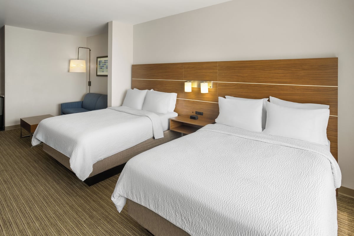 A hotel room with two beds, a desk, a chair, and a wall-mounted lamp.