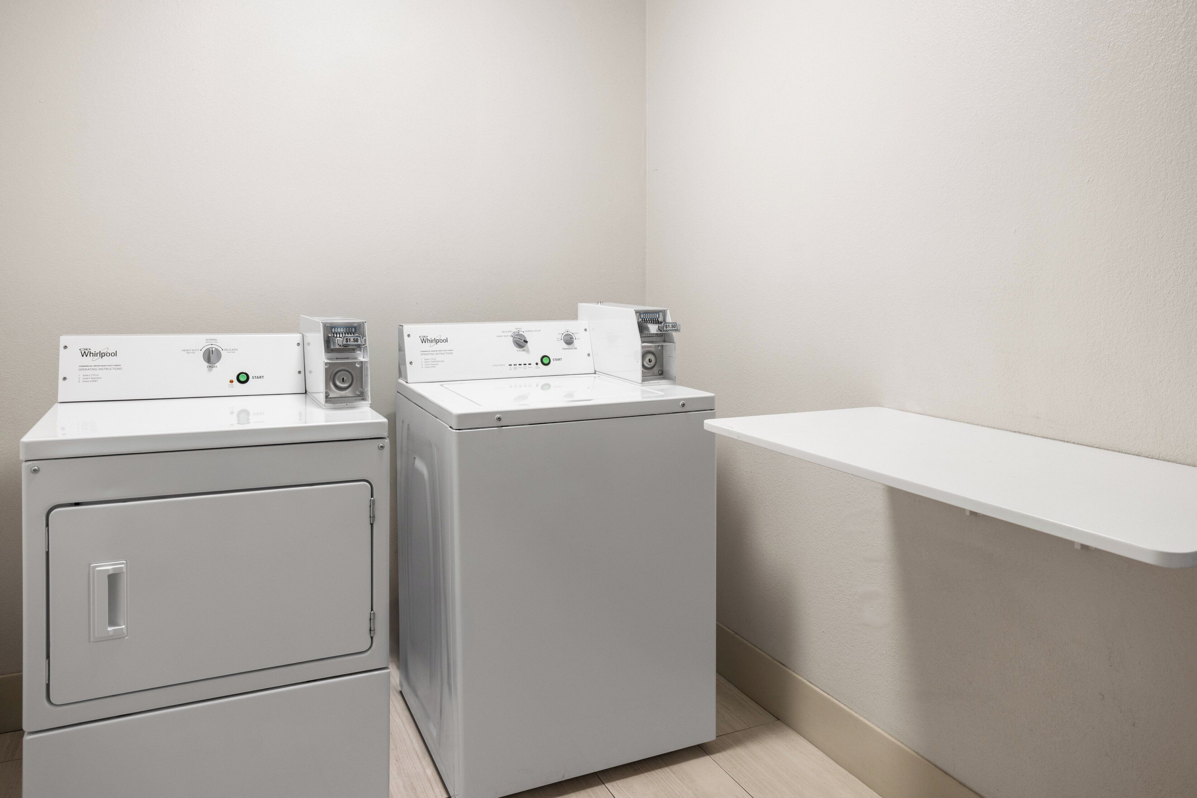 Une buanderie équipée de deux machines à laver et deux sèche-linge superposés, d’une étagère blanche et d’un mur sobre.