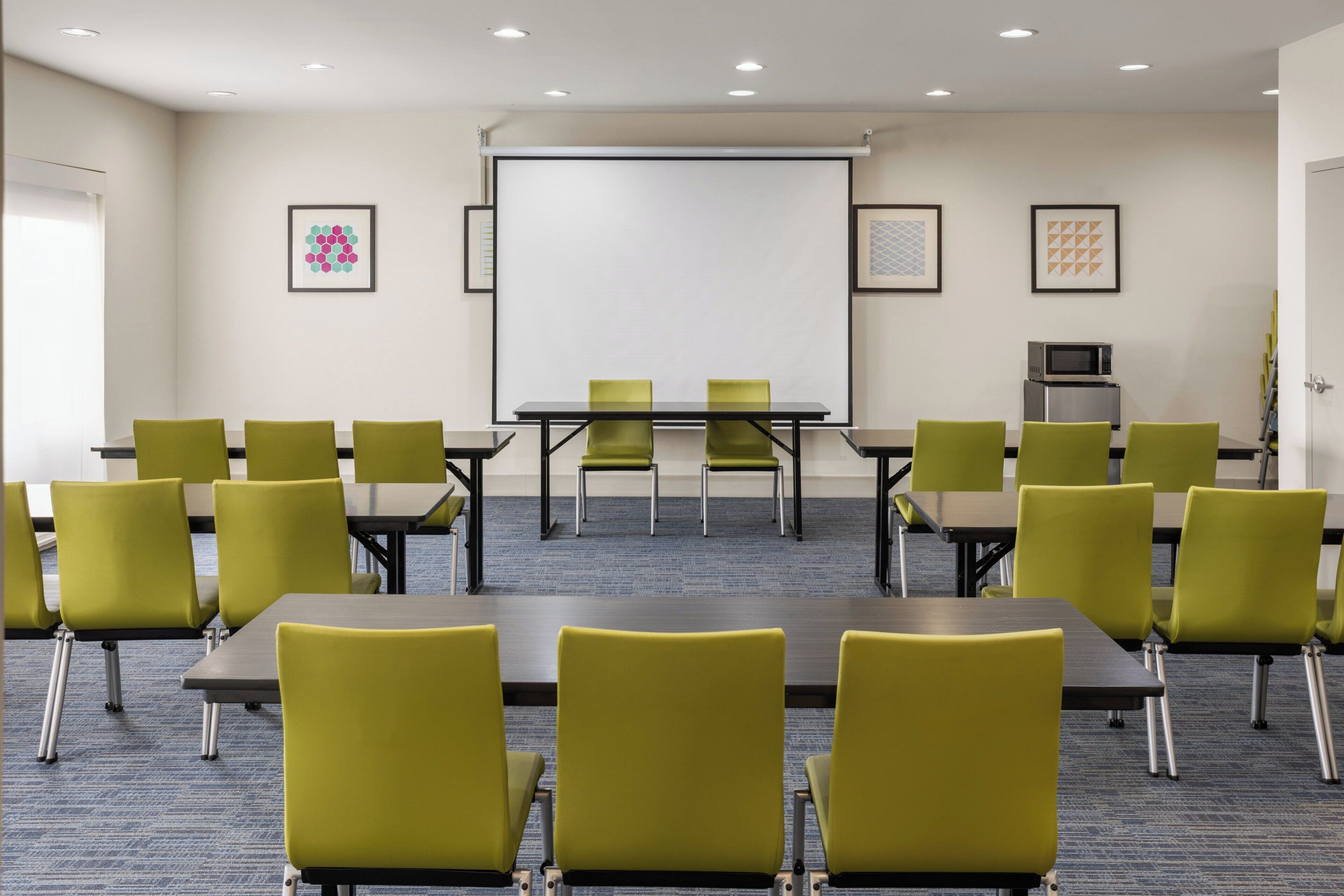 A conference room with a projector screen, green chairs, tables, and a microwave on the wall.